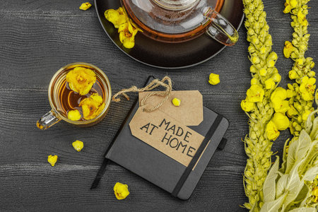 Glass cup of mullein flower tea with teapot and notebook on black wooden table, healthy natural...