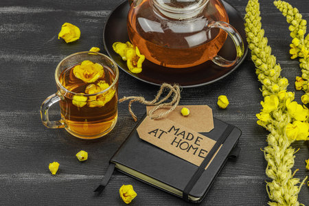 Glass cup of mullein flower tea with teapot and notebook on black wooden table, healthy natural...