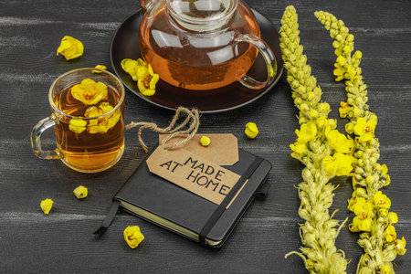 Glass cup of mullein flower tea with teapot and notebook on black wooden table, healthy natural...
