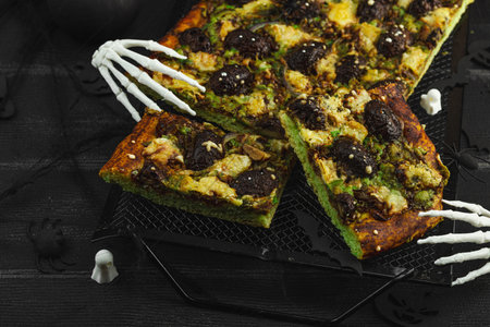 Spooky homemade pizza with green dough, mushroom skulls, and melted cheese. Halloween dark food composition with skeleton hands and black background, close upの写真素材
