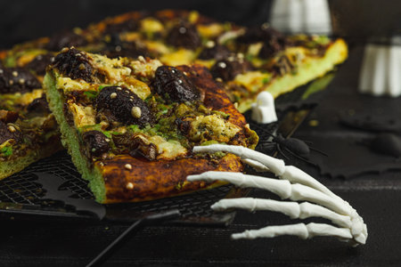Spooky homemade pizza with green dough, mushroom skulls, and melted cheese. Halloween dark food composition with skeleton hands and black background, close upの写真素材