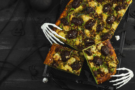 Spooky homemade pizza with green dough, mushroom skulls, and melted cheese. Halloween dark food composition with skeleton hands and black background, top viewの写真素材