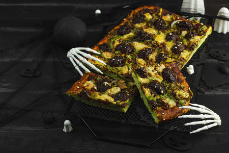 Spooky homemade pizza with green dough, mushroom skulls, and melted cheese. Halloween dark food composition with skeleton hands and black background, copy spaceの写真素材