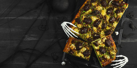 Spooky homemade pizza with green dough, mushroom skulls, and melted cheese. Halloween dark food composition with skeleton hands and black background, banner formatの写真素材