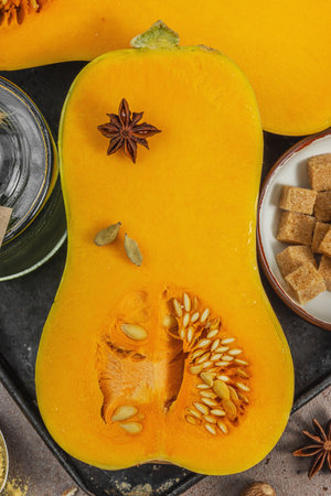 Half butternut squash on a dark tray with baking ingredients. Brown sugar, cinnamon sticks, ginger, nutmeg. Essential spices for traditional autumn pie and dessert recipes, warm brown background, top viewの写真素材