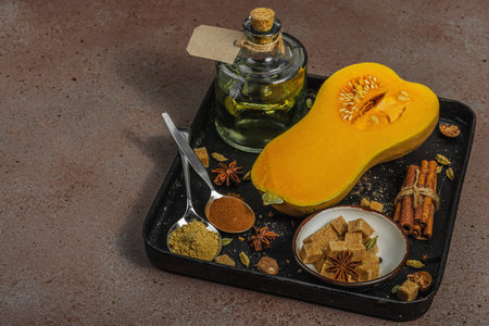 Half butternut squash on a dark tray with baking ingredients. Brown sugar, cinnamon sticks, ginger, nutmeg. Spices for traditional autumn pie and dessert recipes, warm brown background, copy spaceの写真素材