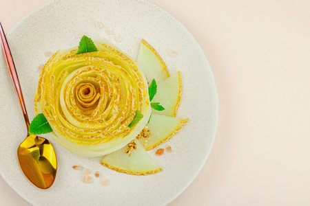 Elegant melon carpaccio dessert shaped like a flower, garnished with mint, spices and pink salt crystals on a ceramic plate. Light peach background, top viewの写真素材