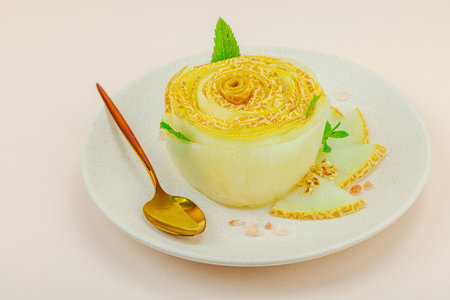 Elegant melon carpaccio dessert shaped like a flower, garnished with mint, spices and salt crystals on a ceramic plate. Light peach background, close upの写真素材