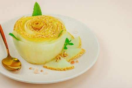 Elegant melon carpaccio dessert shaped like a flower, garnished with mint, spices and pink salt crystals on a ceramic plate. Light peach background, close upの写真素材