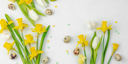 Easter background with yellow daffodils, white tulips, quail eggs, and cute decorative bunnies on a light textured surface. Spring floral arrangement for a holiday greeting card, banner formatの写真素材