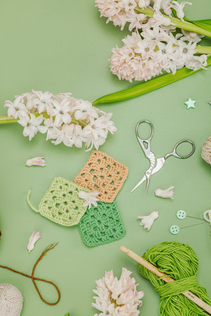 Creative hobby and handmade craft concept. Flat lay of crochet granny squares, yarn skeins, crochet hooks, and pink hyacinth flowers on a green background, top view