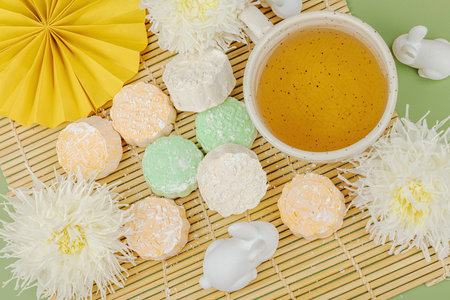 Mid-Autumn Festival flat lay with mooncakes, green tea cup, white rabbits, chrysanthemums, and paper fan on a bamboo mat. Asian traditional celebration concept, top view