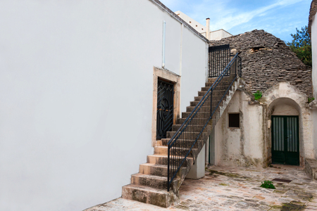 An old stone staircase with iron railing next to the house. Courtyard with a trulli in the city of Alberobello. Brick gray roof, white stone walls with limestone whitewash. City landscape. Free space for text.の写真素材