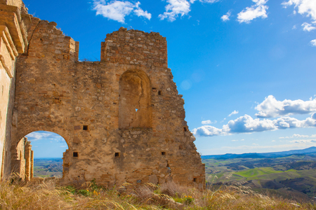 Beautiful asymmetric destroed wall with a hole for passage against the background of green fields, blue sky and white clouds in Craco, Basilicata, Italy. Dry grass, grains and yellow wildflowers.の写真素材