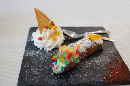 Traditional delicious sicilian cannoli cake. Dessert with waffle, orange, cream, ricotta cheese, marmalade and candied fruit on a stand made of black stone with powdered sugar and descaling spoonsの写真素材