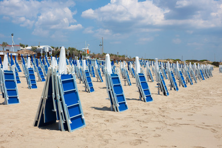 A lot of bright blue closed chaise lounges and beach umbrellas on a sandy beach without people on a blue sky background with white clouds. European holidays in Italyの写真素材