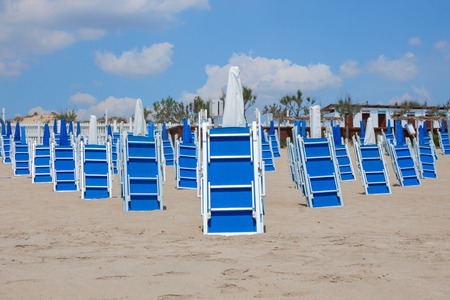 A lot of bright blue closed chaise lounges and beach umbrellas on a sandy beach without people on a blue sky background with white clouds. European holidays in Italyの写真素材