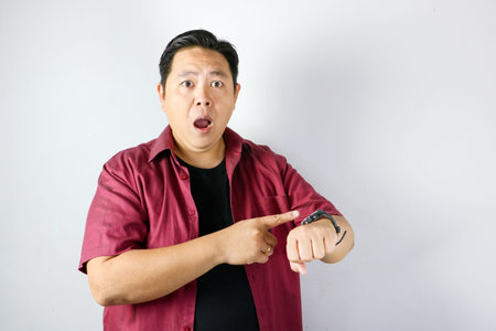 Shocked Asian man pointing finger at his watch on white background
