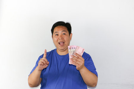 Young asian man holding money and pointing up on white background.