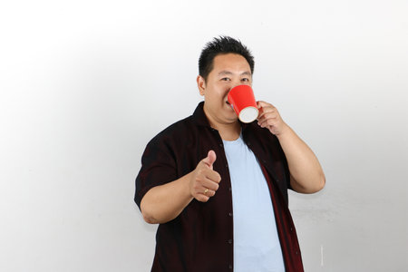 Asian man drinking from a cup and showing thumbs up sign on white background.の写真素材