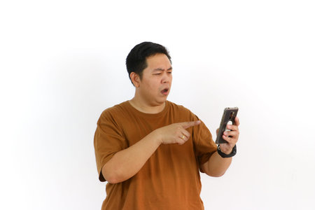 A curious Asian man looking confused or uncertain while using his mobile phone on a white background.の写真素材