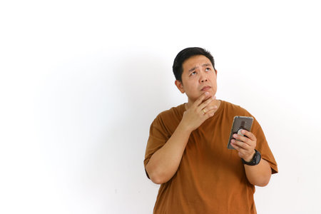 A curious Asian man looking confused or uncertain while using his mobile phone on a white...