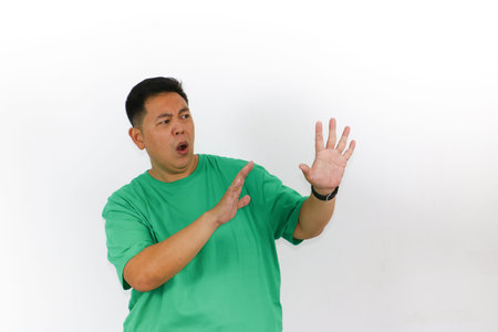 An Asian man in a green t-shirt strongly refusing a request or an offer, using both hands in a defensive rejection gesture.の写真素材