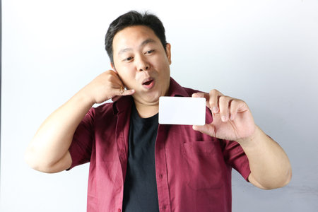An Asian man with an open mouth makes a call me gesture with his right hand while holding a blank...