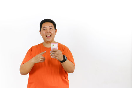 Excited man in orange t-shirt celebrates financial success, holding Indonesian Rupiah banknotes with...