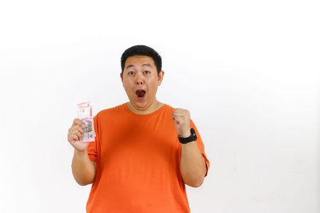 Excited man in orange t-shirt celebrates financial success, holding Indonesian Rupiah banknotes with joyful expression on white background.の写真素材
