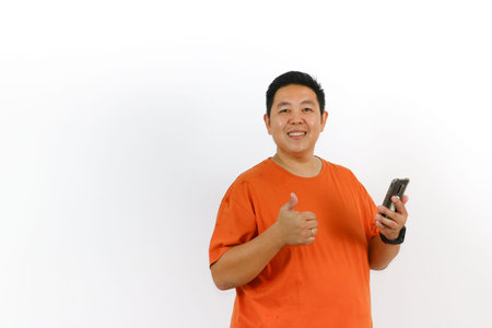 Excited Asian man in orange t-shirt using smartphone and giving thumbs up sign, expressing...