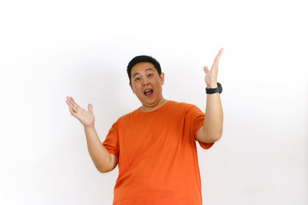 Amazed Asian man in orange t-shirt reacts with open mouth and raised hands, expressing total disbelief and shock.の写真素材