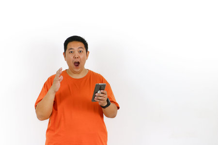 Amazed Asian man in orange t-shirt holding smartphone reacts with open mouth and raised hand, expressing total shock.の写真素材