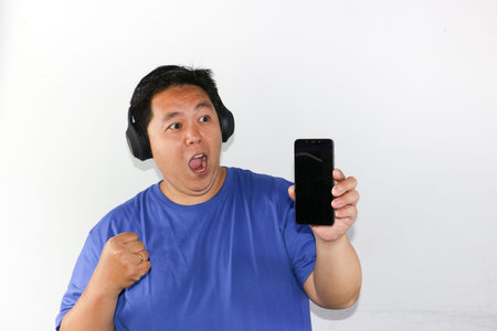 Portrait of a happy Indonesian guy celebrating victory while holding a smartphone and wearing...