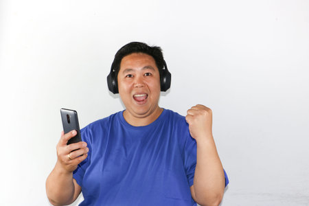 Portrait of a happy Indonesian guy celebrating victory while holding a smartphone and wearing headphonesの写真素材