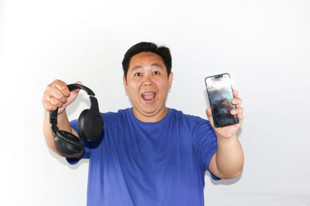 Portrait of Indonesian man holding a smartphone and black headphones with a joyful expressionの写真素材