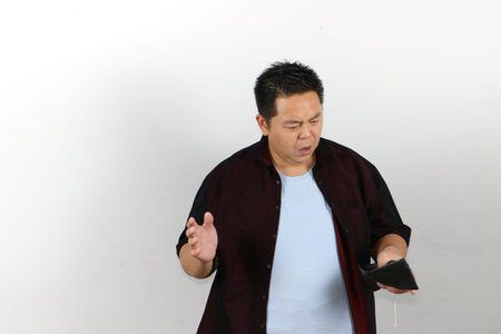 An Asian man expresses deep shock while looking into his completely empty black leather wallet.