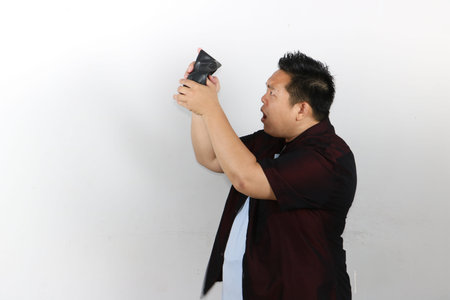 An Asian man expresses deep shock while looking into his completely empty black leather wallet.の写真素材