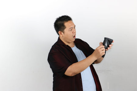 An Asian man expresses deep shock while looking into his completely empty black leather wallet.の写真素材