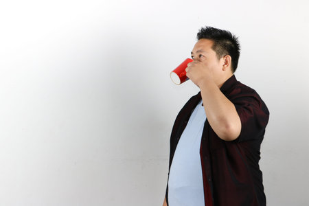 Portrait of Asian man drinking from a red mug against a isolated white background in studioの写真素材