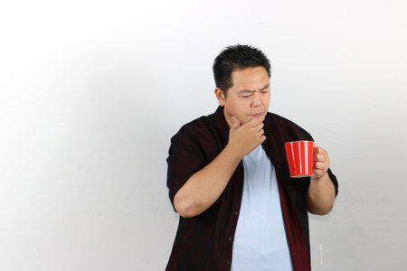 Confused Asian man in casual clothes looking at a red mug while touching his chin on white backgroundの写真素材