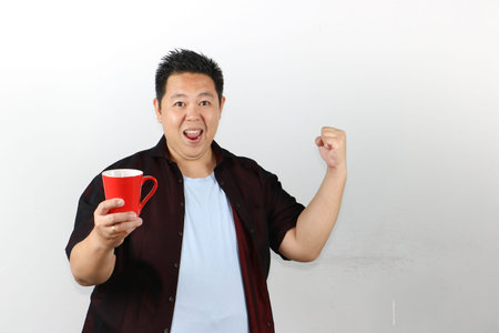 Excited Asian man celebrating success, winning gesture while holding a red mug
