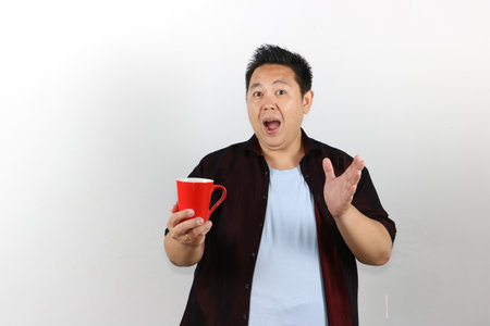 Shocked Asian man in casual clothes pointing at a red mug with open mouth on white backgroundの写真素材