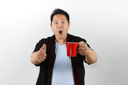 Shocked Asian man in casual clothes pointing at a red mug with open mouth on white background