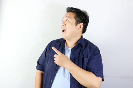 Surprised Indonesian man in blue shirt pointing finger up having a great idea isolated on white...