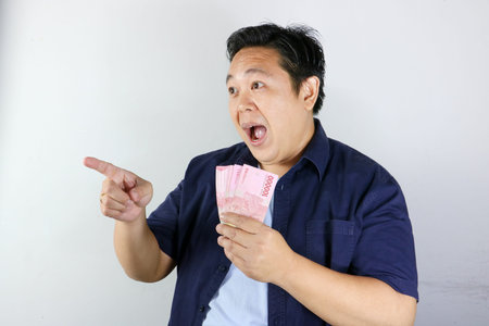 Happy Indonesian man holding cash and pointing finger up having a brilliant financial idea on white...