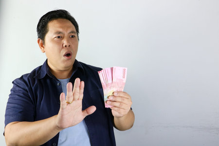 Shocked Indonesian man in blue shirt holding money and making a stop gesture isolated on white...