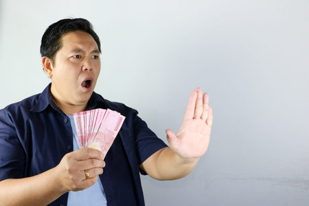 Shocked Indonesian man in blue shirt holding money and making a stop gesture isolated on white...