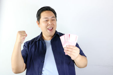 Joyful Southeast Asian man holding rupiah money and raising a fist in victory, winning gesture