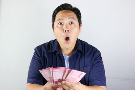 Shocked wow asian man looking at a fan of hundred thousand rupiah bills in his hands